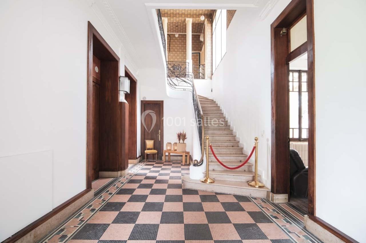 Hall lumineux avec sol en carreaux noirs et roses, escalier en marbre, rambarde en fer forgé et portes en bois.