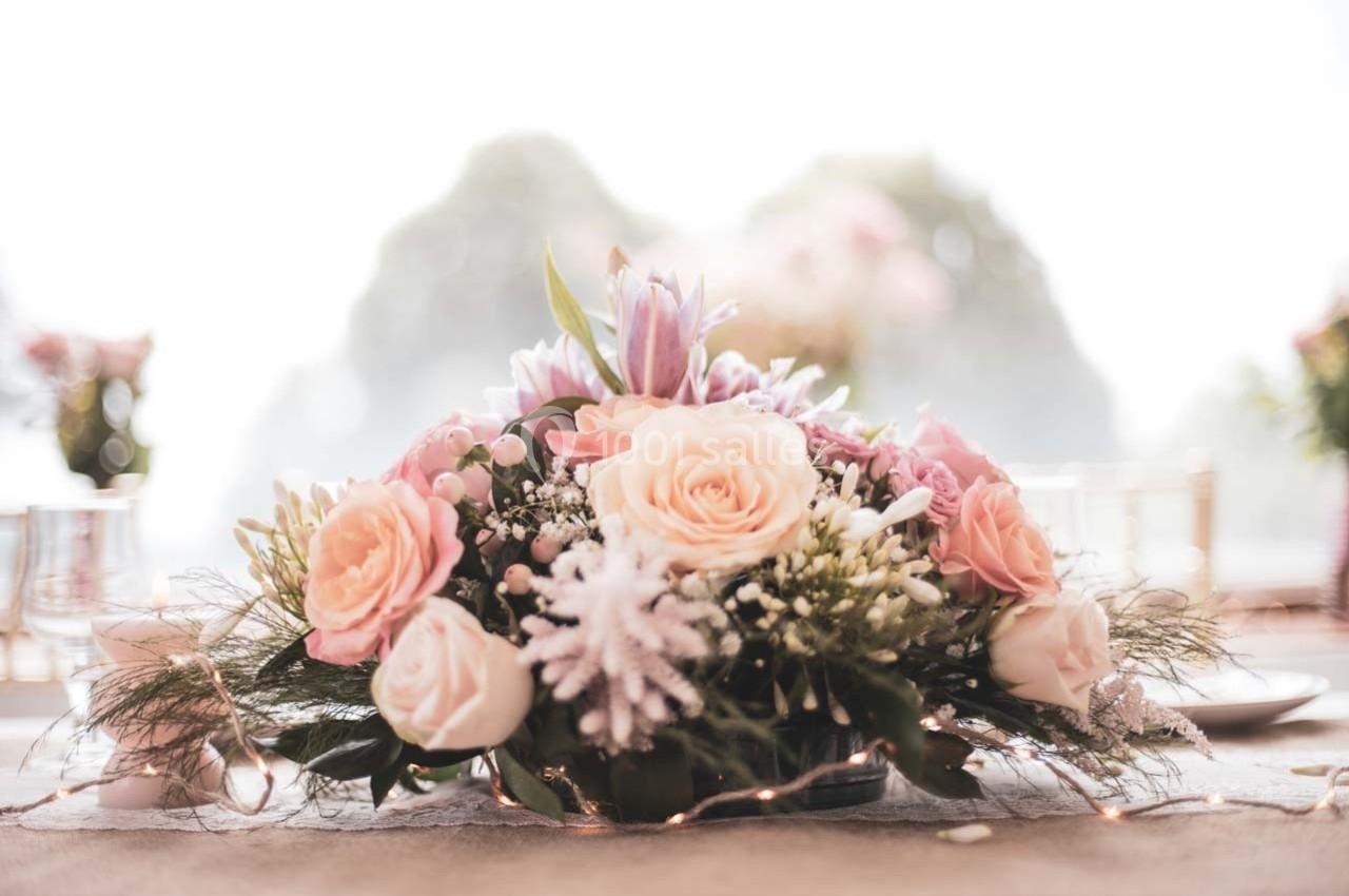 Arrangement floral avec des roses roses, des fleurs blanches et violettes, posé sur une table décorée de guirlandes…