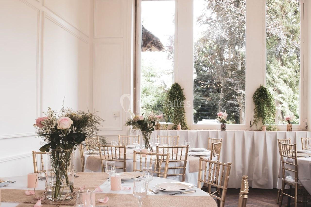 Salle de réception lumineuse avec tables décorées de fleurs, nappes blanches et chaises dorées, vue sur un jardin.