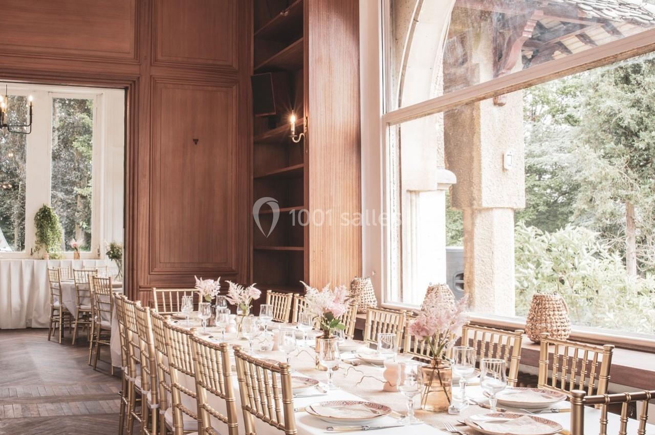Salle de réception élégante avec tables dressées, chaises dorées et grandes fenêtres donnant sur un jardin.