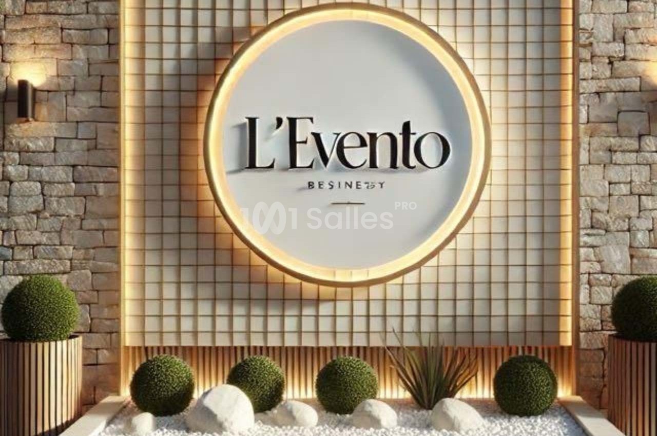Mur en pierre avec un logo circulaire ’L'Evento’ éclairé, entouré de plantes et de galets blancs.
