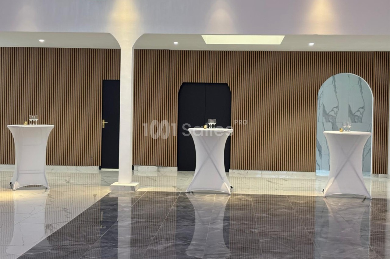 Trois tables hautes recouvertes de nappes blanches sont disposées dans une salle avec un mur à lamelles en bois.
