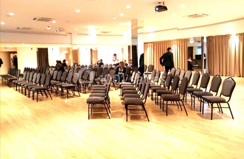 Salle lumineuse avec des rangées de chaises vides alignées, quelques personnes debout à l'arrière-plan.