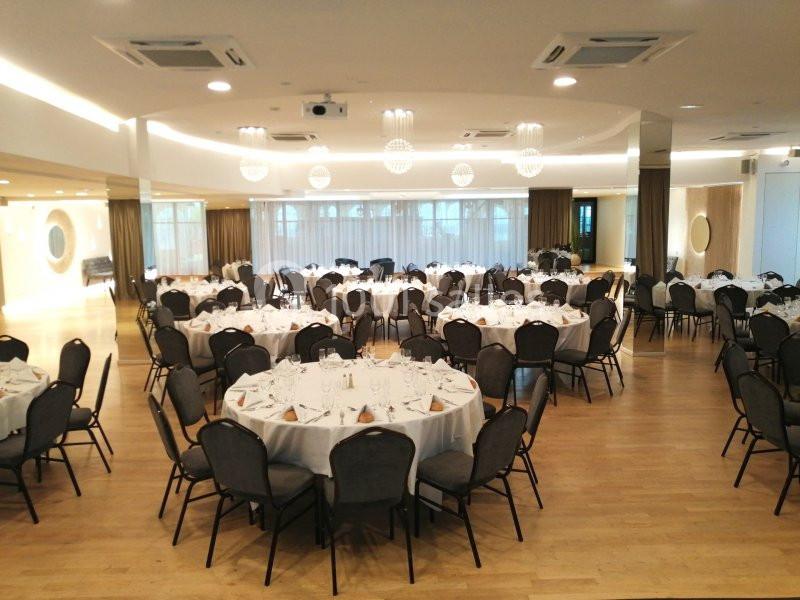 Salle de réception lumineuse avec tables rondes dressées, chaises noires et lustres modernes au plafond.