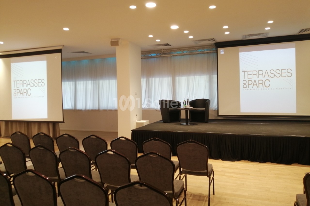 Salle de conférence avec des chaises alignées, une scène équipée de fauteuils et des écrans affichant ’Terrasses du Parc’.