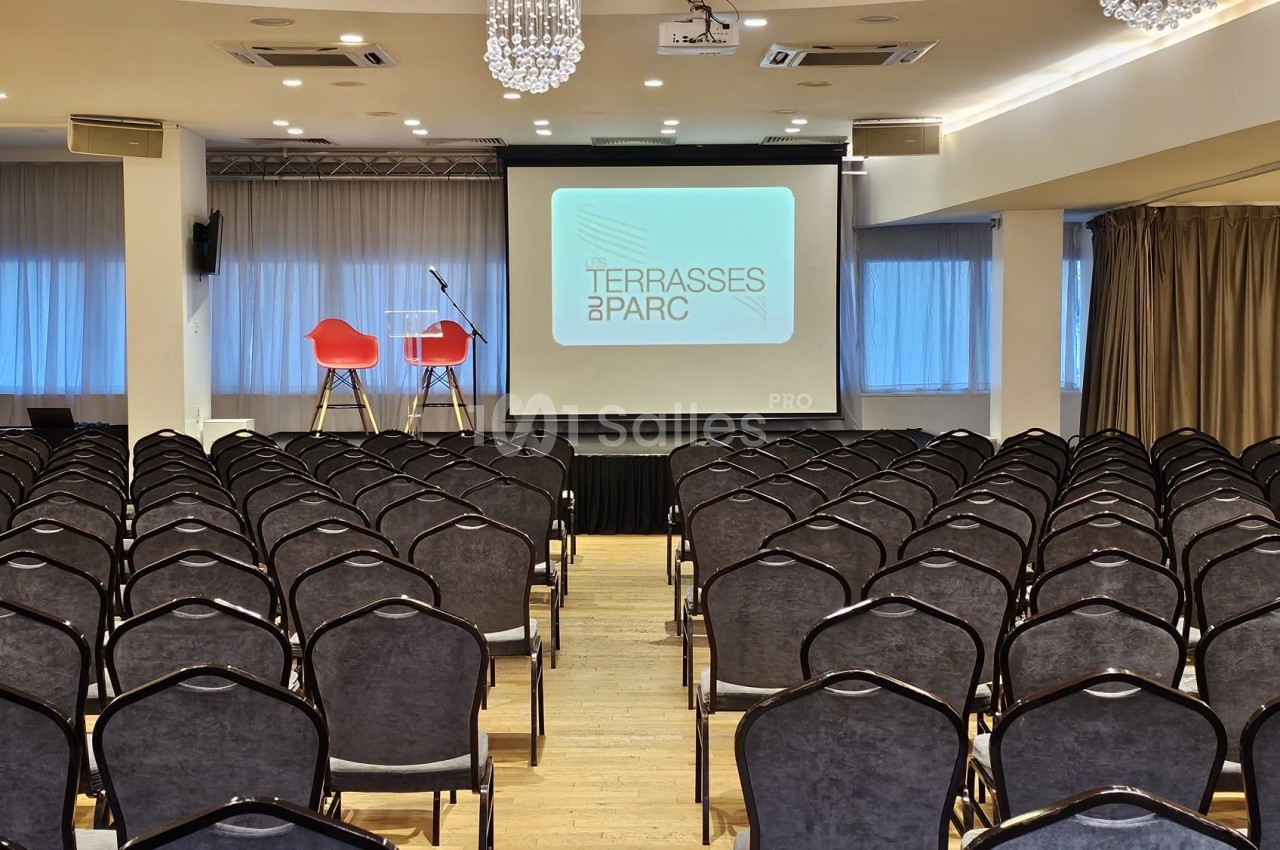 Salle de conférence avec rangées de chaises alignées, une scène équipée de deux chaises et un écran de projection.