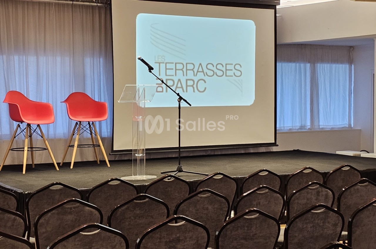 Salle de conférence avec chaises alignées, scène équipée d'un pupitre, micro et écran affichant ’Les Terrasses du Parc’.