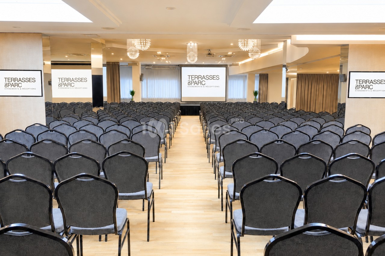 Salle de conférence avec rangées de chaises noires alignées face à deux écrans de projection et un espace lumineux.