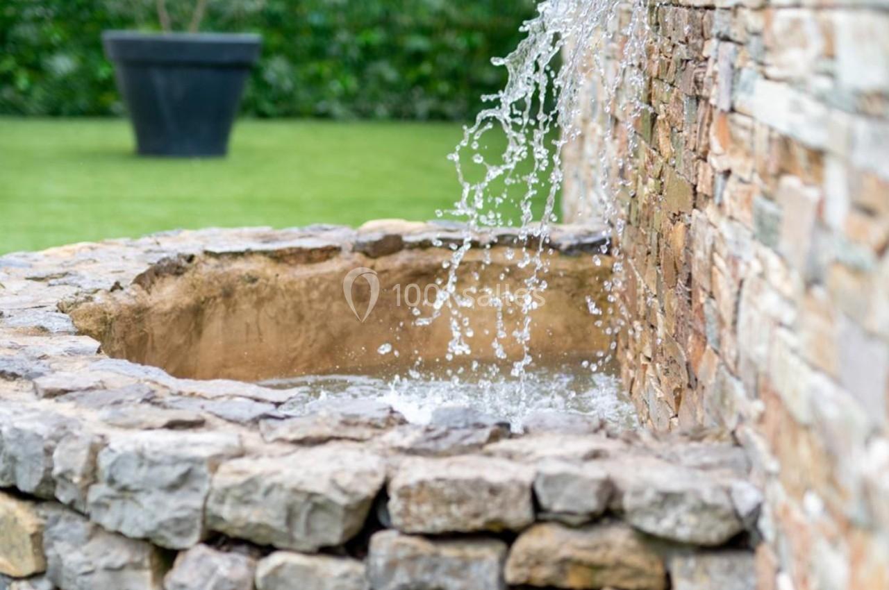 Jet d'eau tombant dans un bassin en pierre, avec un mur en pierre et un jardin verdoyant en arrière-plan.