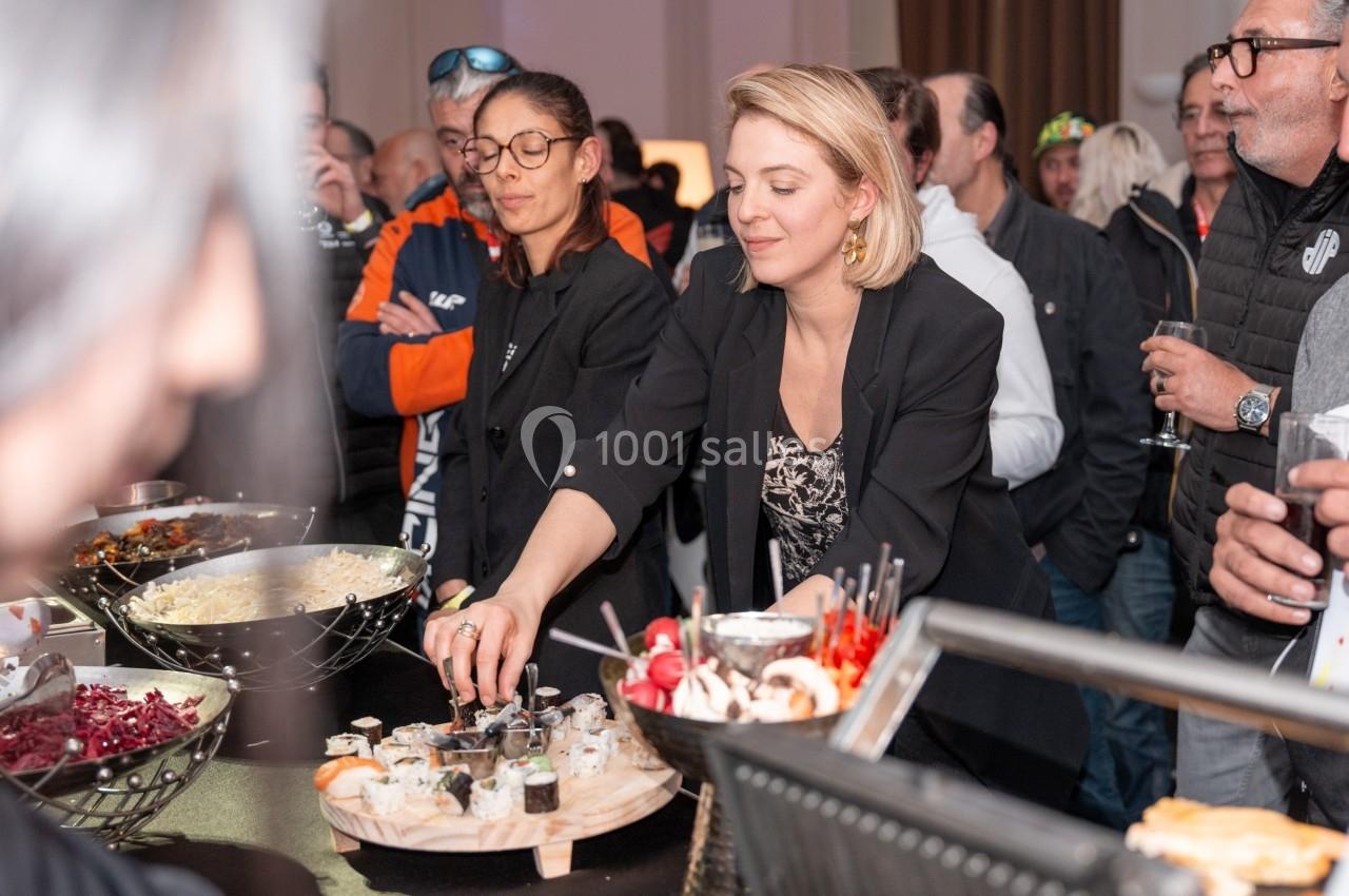 Une femme sert des amuse-bouches sur un buffet lors d'un événement avec plusieurs personnes en arrière-plan.