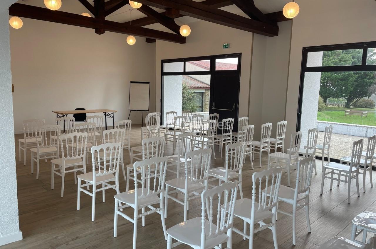 Salle lumineuse avec des chaises blanches alignées, un tableau de conférence et une table, vue sur un espace extérieur.