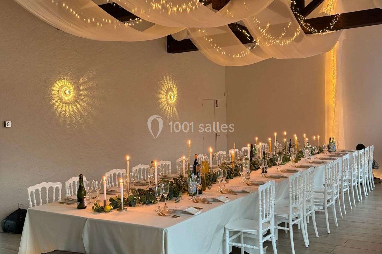 Grande table de banquet décorée avec des bougies, guirlandes lumineuses et nappes blanches dans une salle avec poutres…