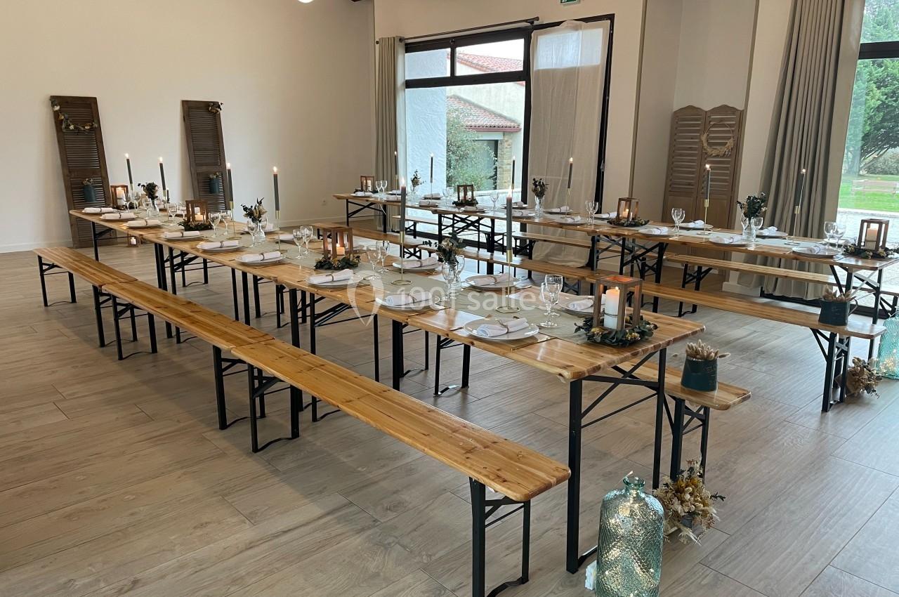 Salle décorée pour un repas, avec de longues tables en bois, bancs assortis, bougies et vaisselle élégante.
