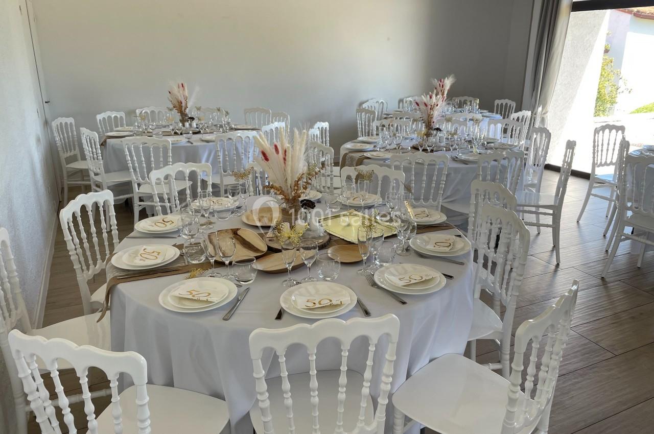 Salle décorée pour un événement avec des tables rondes dressées, nappes blanches, vaisselle élégante et centres de table…