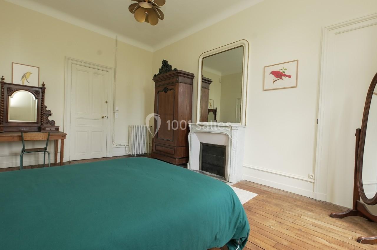 Chambre lumineuse avec lit à couverture verte, armoire en bois, cheminée, miroir et parquet au sol.