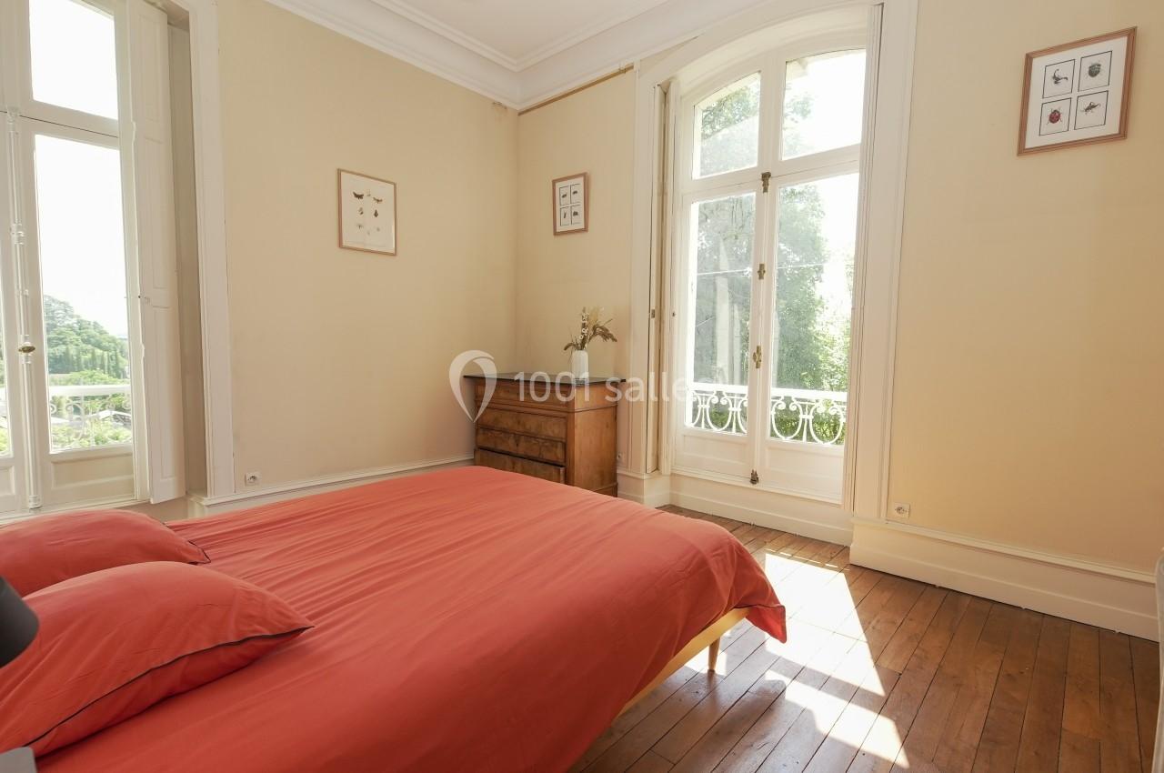 Chambre lumineuse avec lit à draps rouges, parquet en bois, commode, grande fenêtre et vue sur un jardin verdoyant.