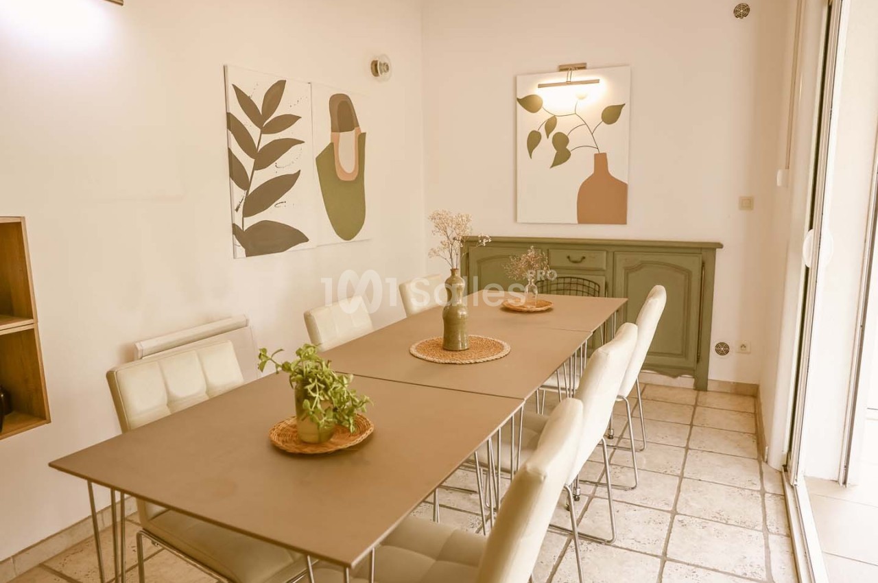 Salle à manger lumineuse avec une grande table, des chaises blanches, des décorations végétales et des tableaux modernes.