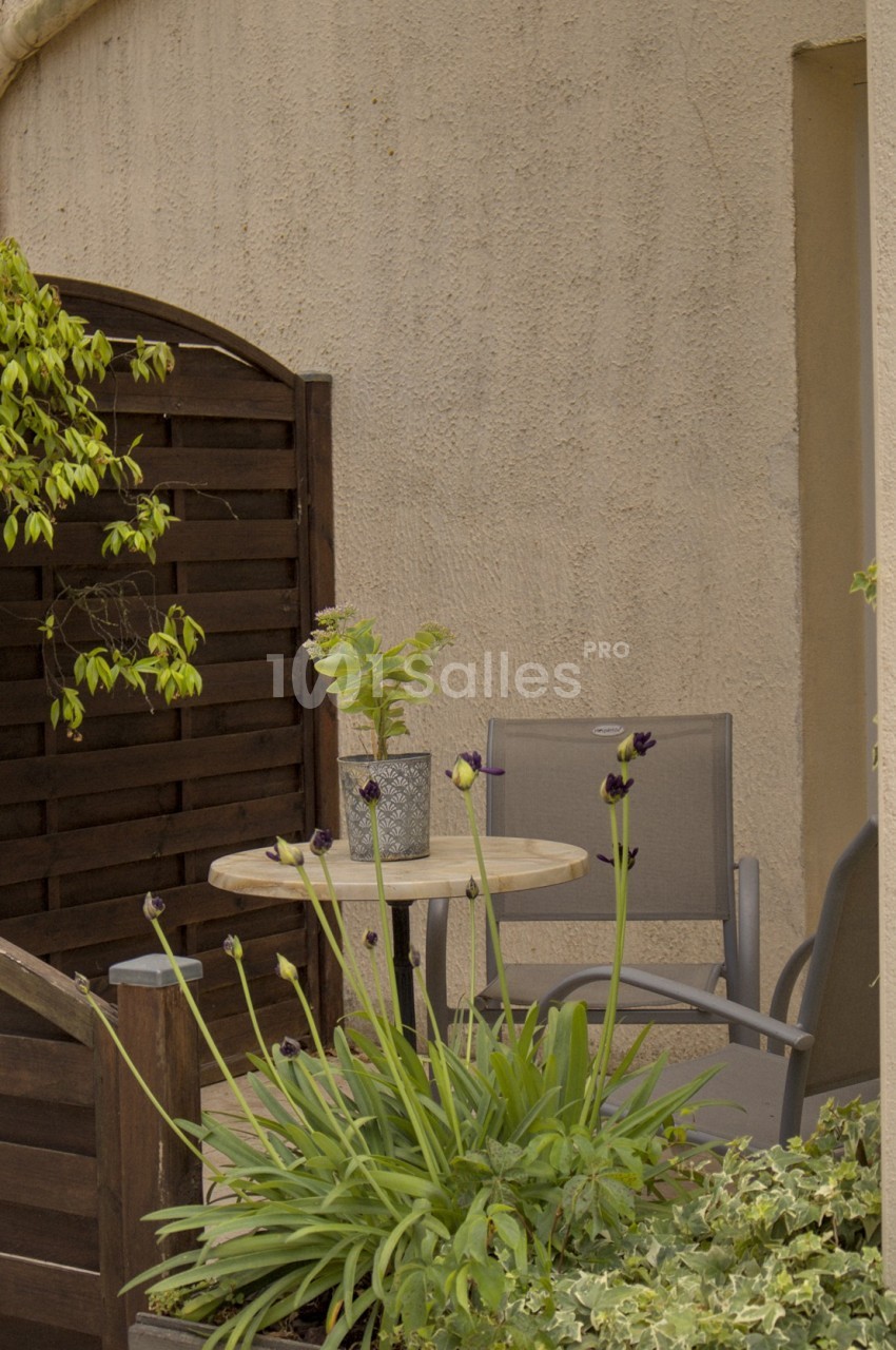 Petit coin de terrasse avec table ronde, deux chaises, plantes vertes et fleurs devant un mur beige.