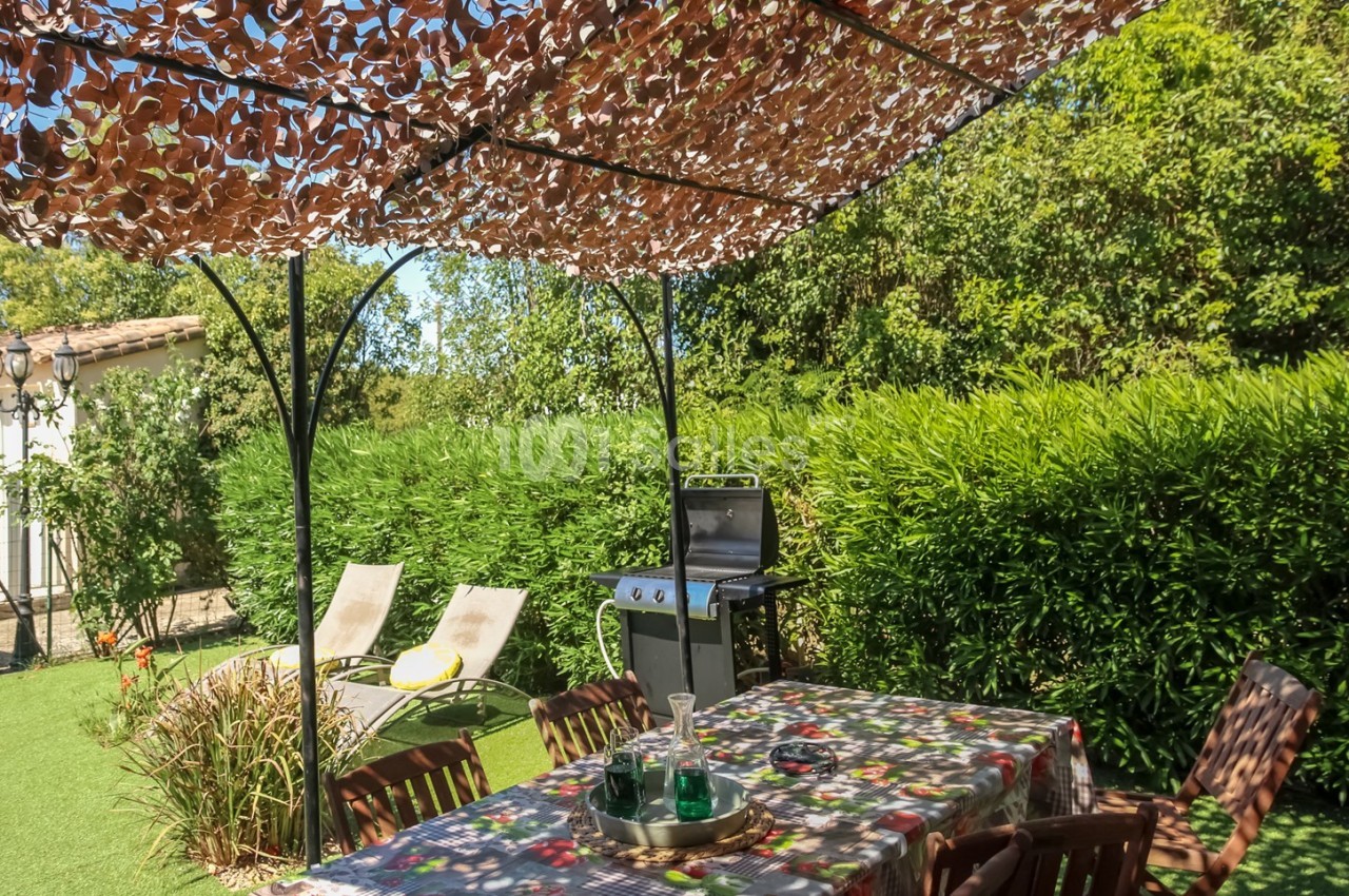 Table de jardin sous une tonnelle ombragée, entourée de verdure, avec un barbecue et des transats en arrière-plan.