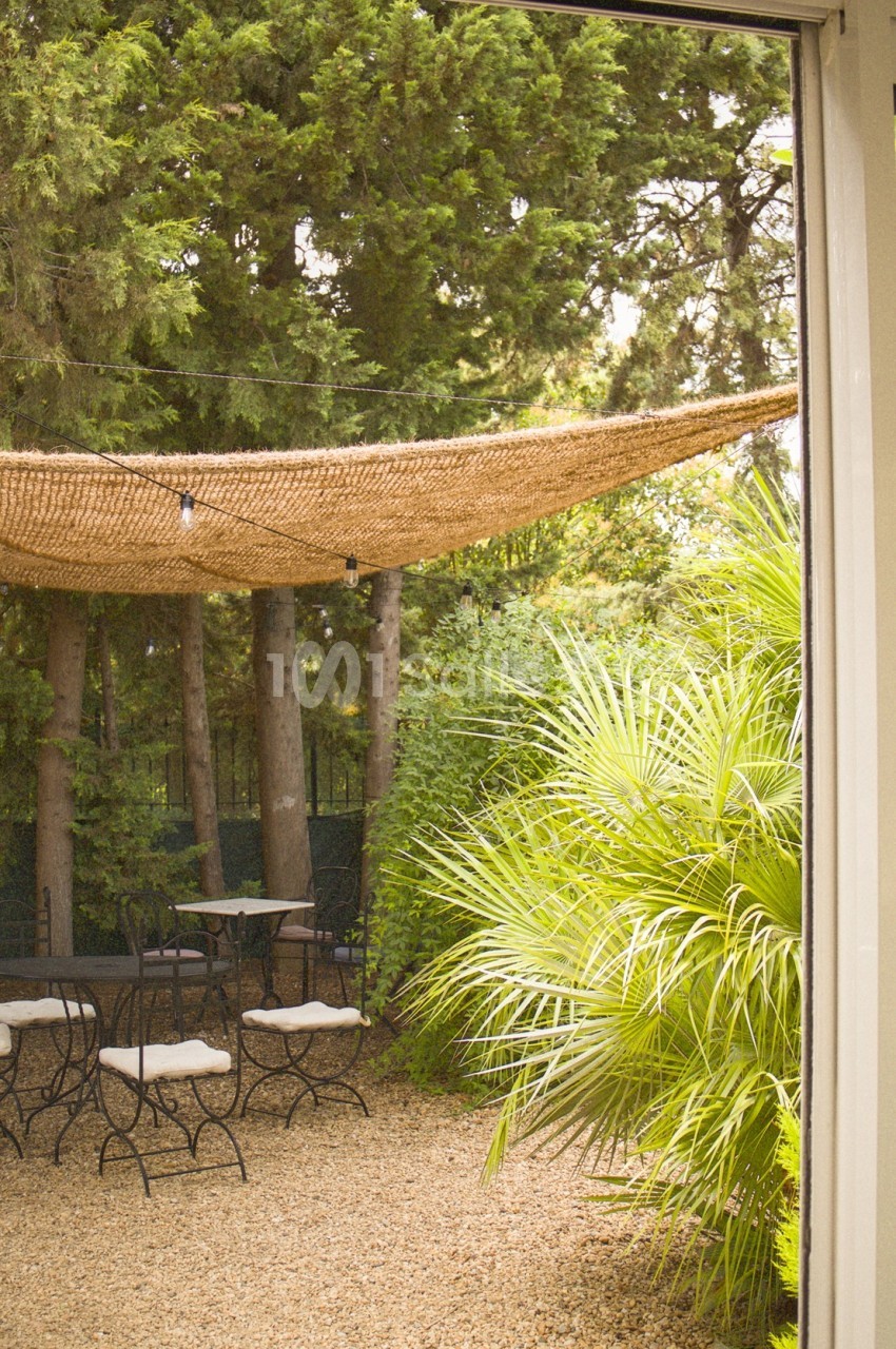 Vue d'une terrasse extérieure avec des tables et chaises, ombragée par une toile, entourée de végétation dense.