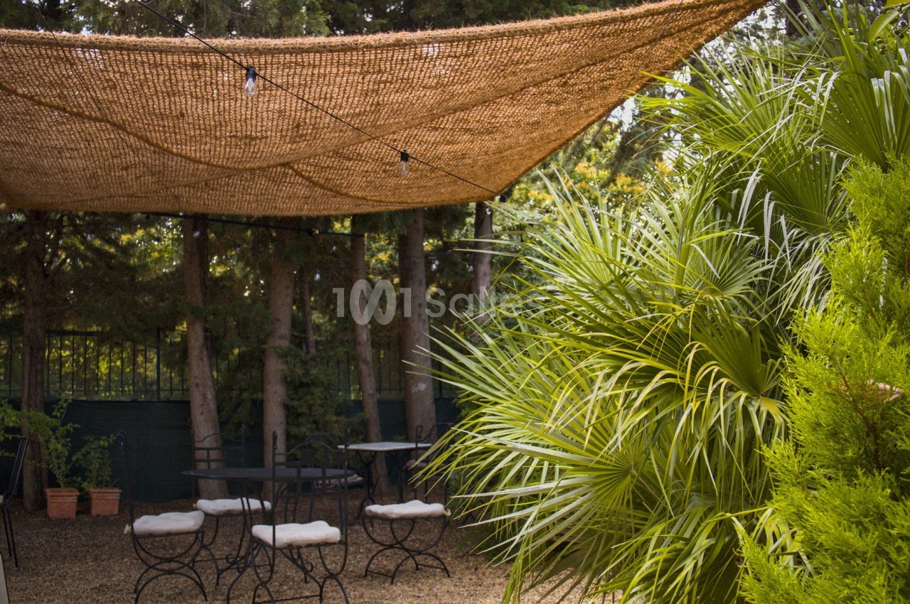 Terrasse extérieure ombragée avec des tables, des chaises et une végétation dense sous une toile en fibre naturelle.