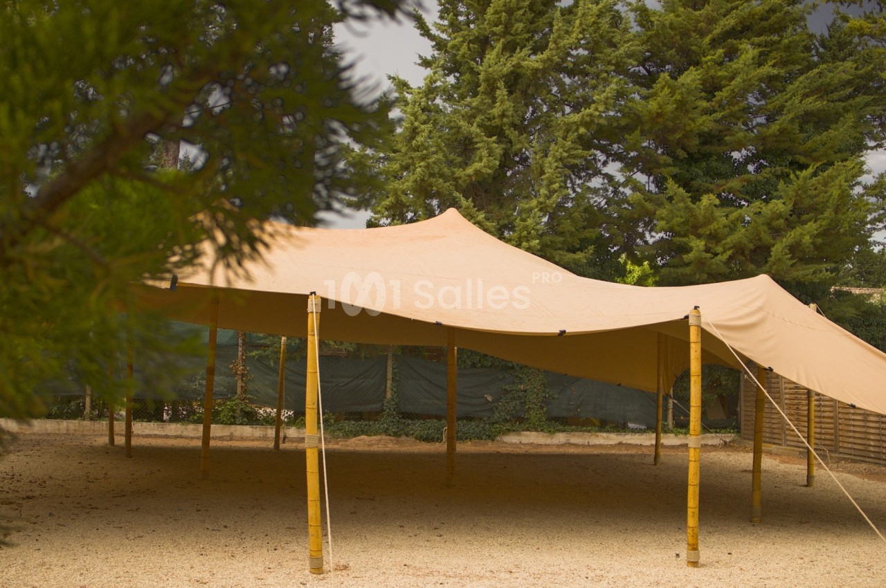 Tente beige tendue sur des poteaux en bambou, installée sur un sol gravillonné entouré d'arbres.