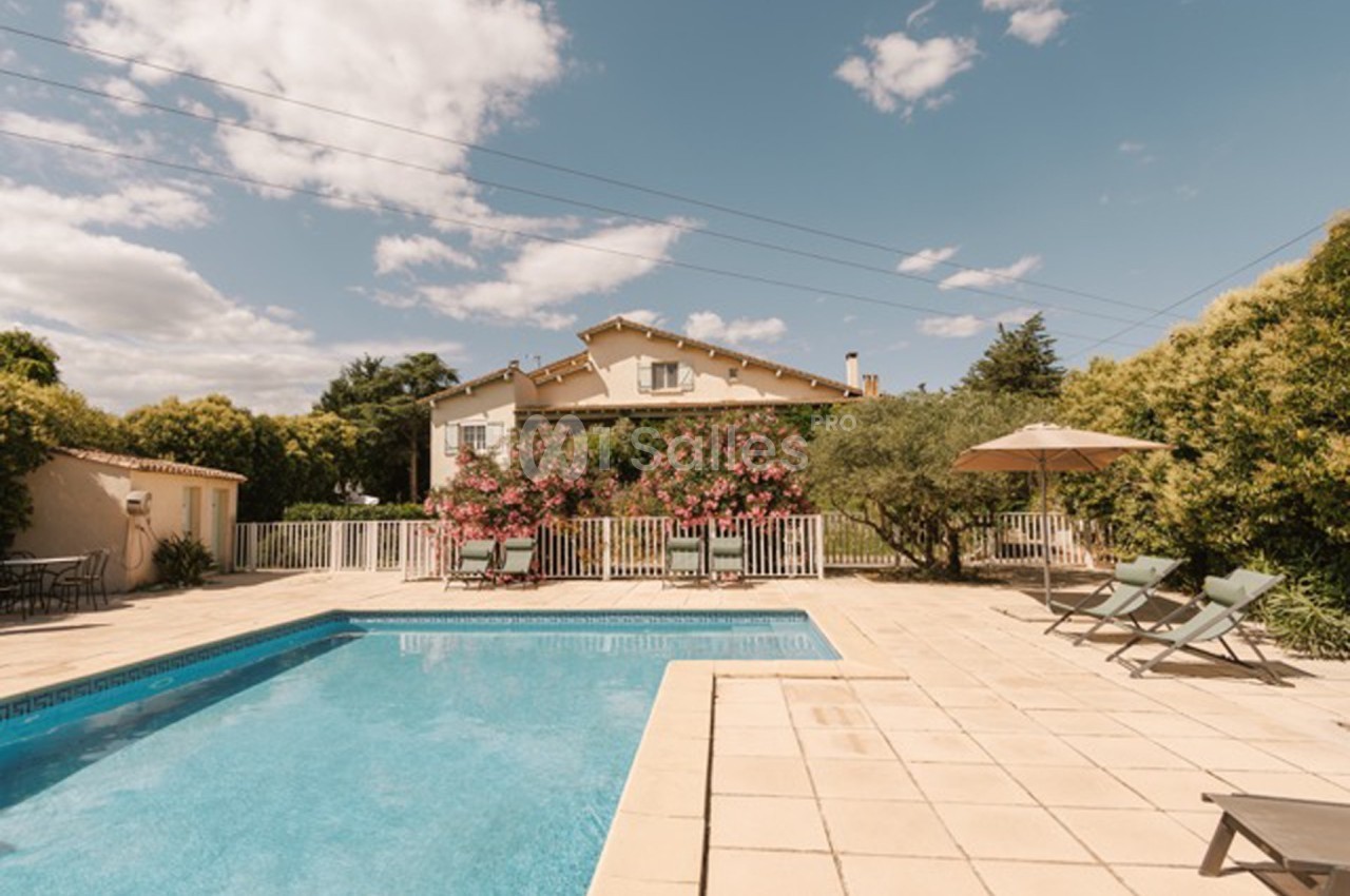 Maison avec jardin, piscine, terrasse carrelée, chaises longues et parasol, entourée de végétation sous un ciel dégagé.