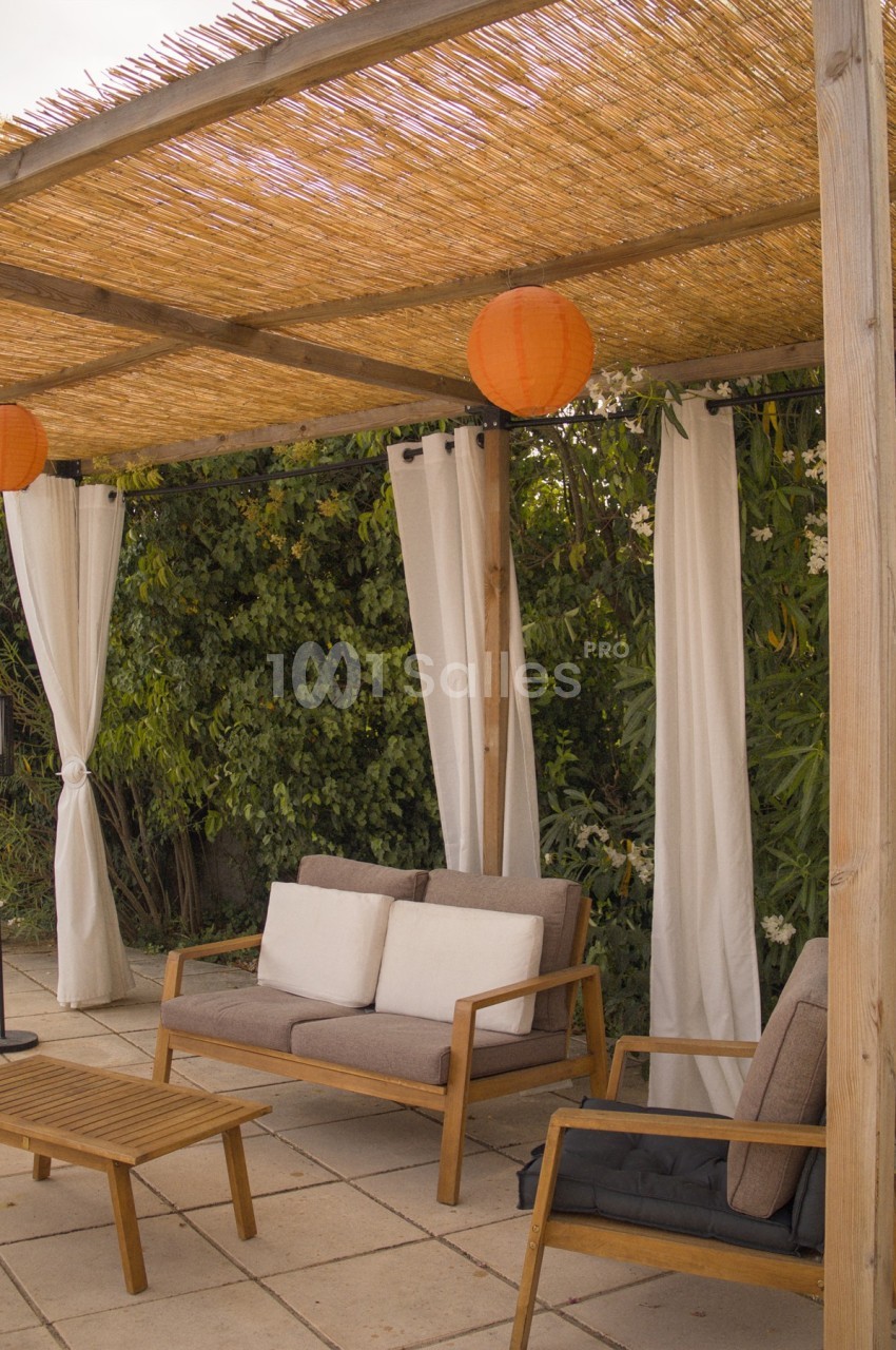 Salon de jardin en bois sous une pergola avec toit en roseaux, lanternes orange et rideaux blancs, entouré de verdure.