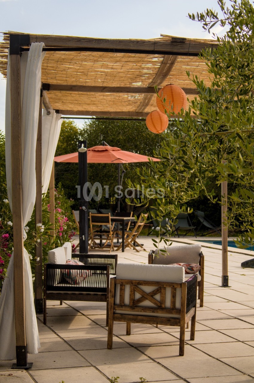 Pergola en bois avec rideaux blancs, mobilier de jardin, lanternes suspendues et parasol près d'une piscine.