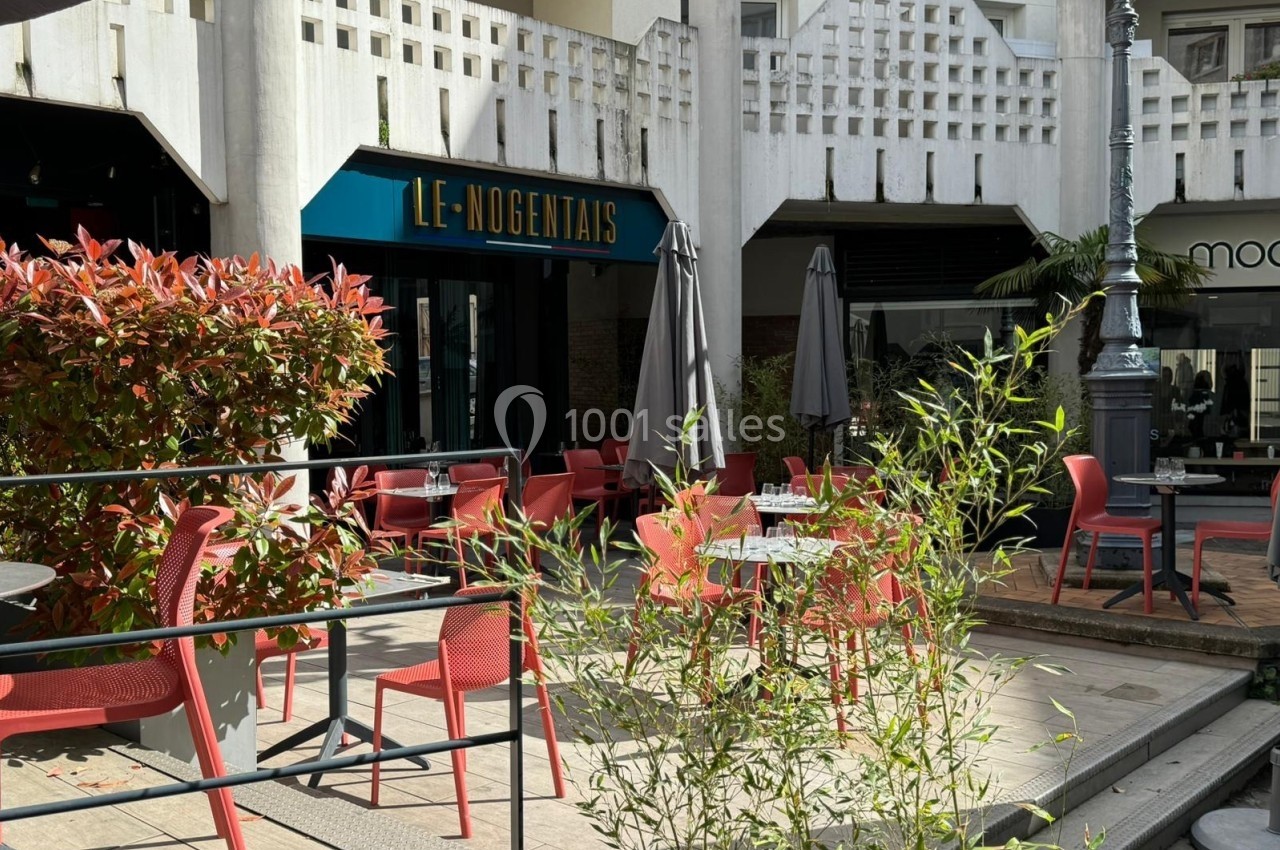 Terrasse d'un café avec des chaises rouges, des parasols fermés et des plantes, située devant un bâtiment moderne.