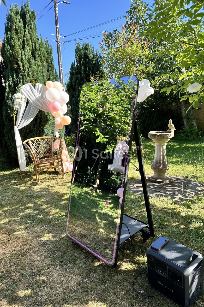 Miroir sur pied dans un jardin ensoleillé, entouré de ballons, d'arbres et d'une décoration festive.