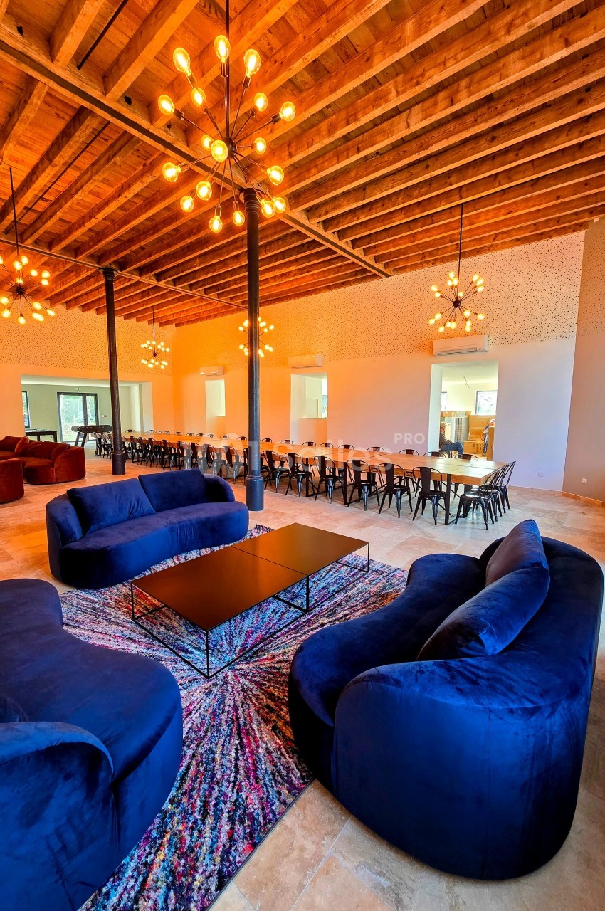 Salle spacieuse avec canapés bleus, table longue entourée de chaises, plafond en bois et luminaires suspendus.