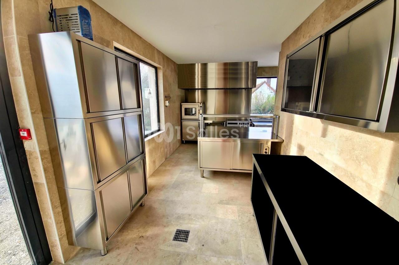 Cuisine professionnelle moderne avec équipements en acier inoxydable, murs en pierre beige et lumière naturelle.