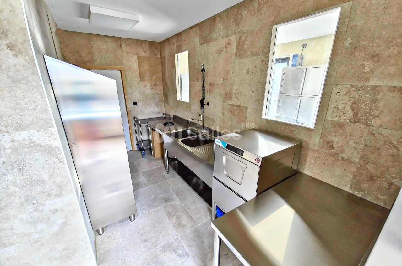 Cuisine professionnelle avec évier inox, lave-vaisselle, plan de travail et placard métallique, murs en carrelage beige.