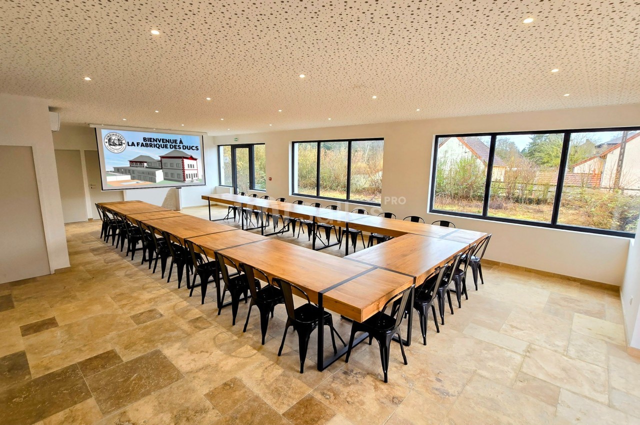 Salle de réunion lumineuse avec de grandes fenêtres, tables en bois disposées en U et chaises noires.