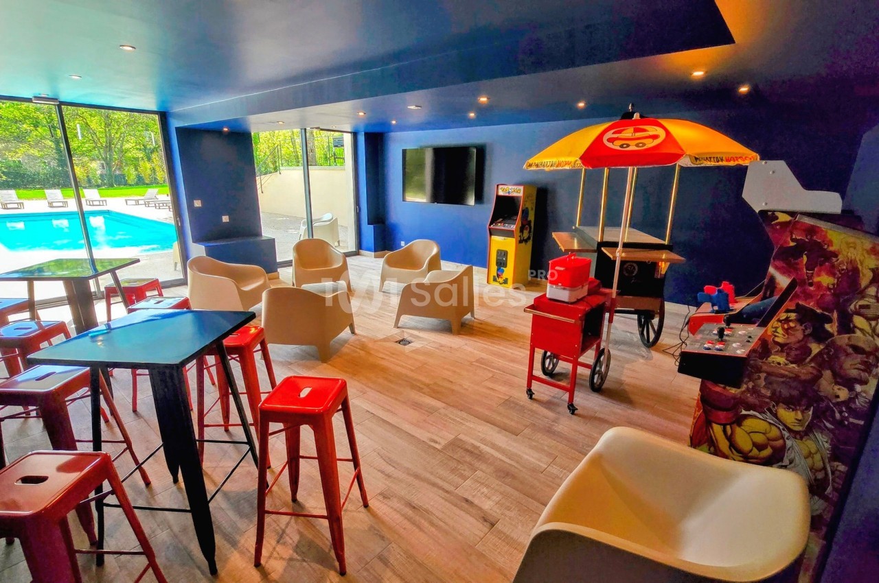 Salle de loisirs colorée avec jeux d'arcade, chaises modernes, tables hautes et vue sur une piscine extérieure.