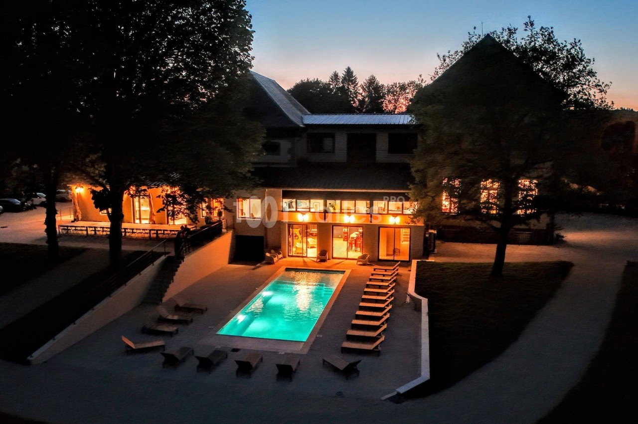 Vue nocturne d'une maison éclairée avec piscine extérieure entourée de chaises longues et d'arbres.