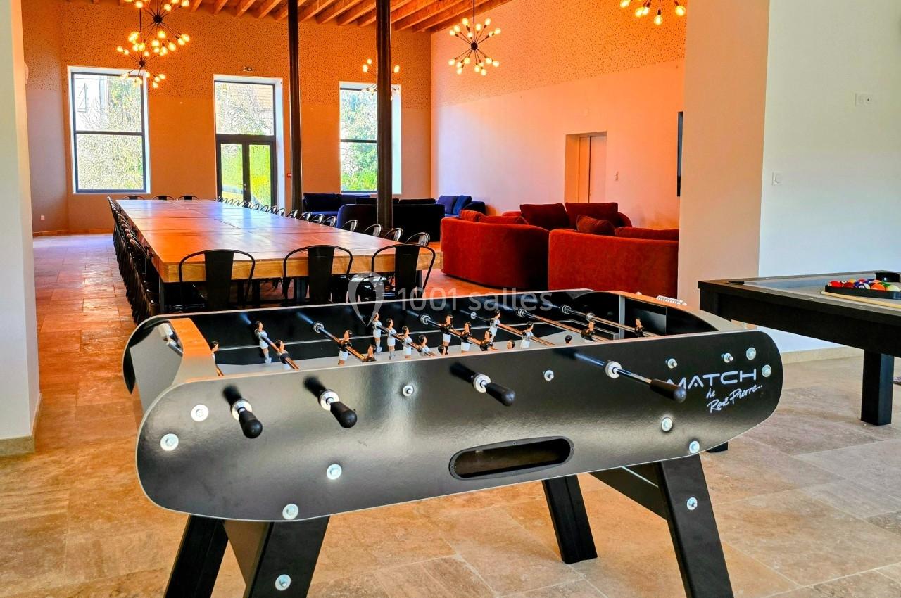 Baby-foot noir au premier plan dans une grande salle lumineuse avec une longue table, des canapés et un billard.