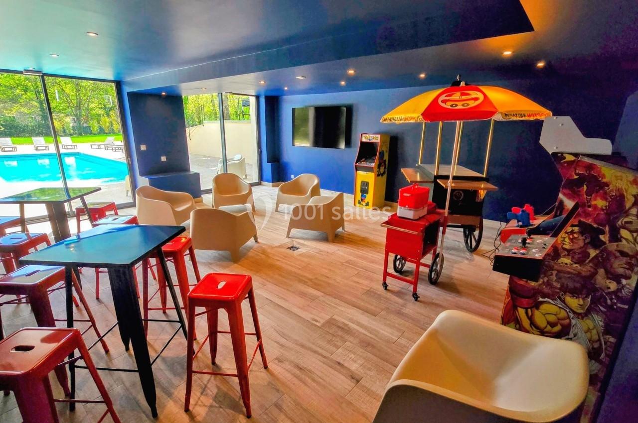 Salle de loisirs colorée avec jeux d'arcade, chaises modernes, tables hautes et vue sur une piscine extérieure.