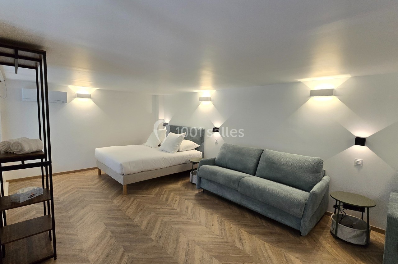 Chambre lumineuse avec lit double, canapé, parquet en chevrons et étagère en métal, éclairée par des appliques murales.