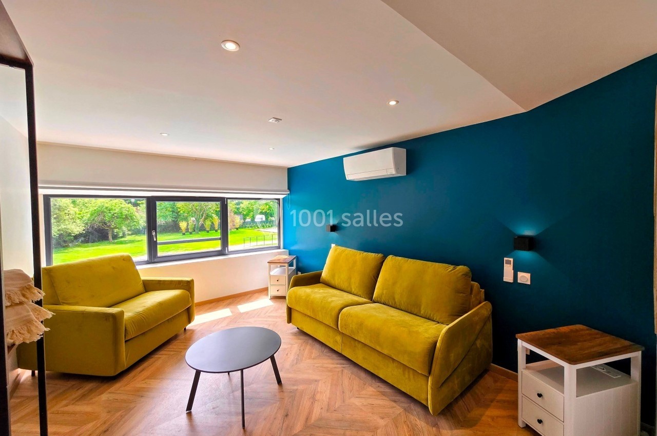 Salon lumineux avec deux canapés jaunes, table basse noire, mur bleu, parquet en chevrons et vue sur un jardin.