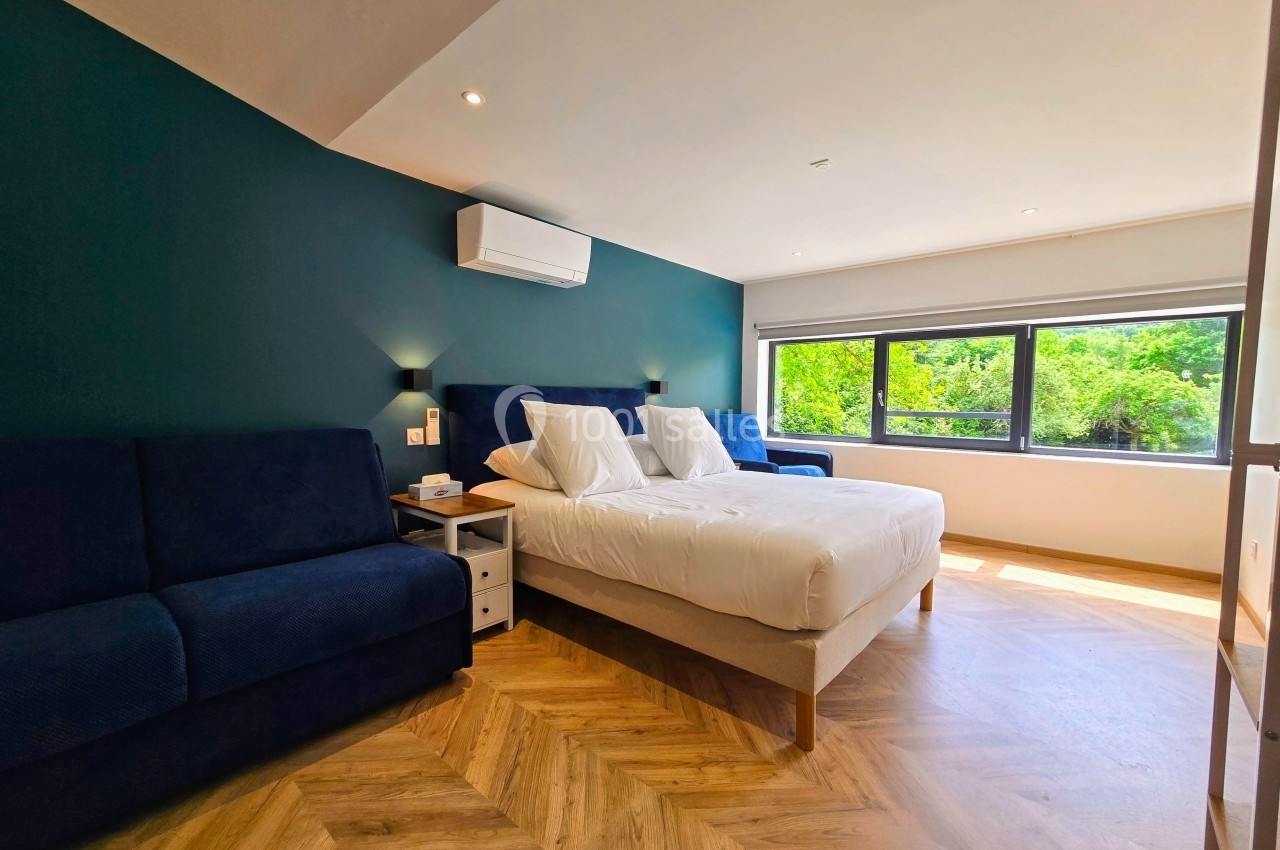 Chambre lumineuse avec lit double, canapé bleu, parquet en bois et grande fenêtre donnant sur un espace vert.