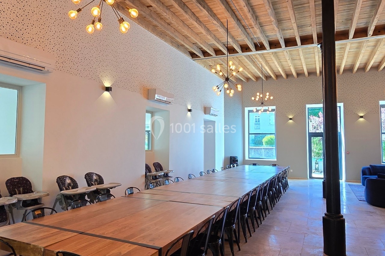 Grande salle lumineuse avec une longue table en bois, des chaises noires, un plafond en bois et des luminaires suspendus.