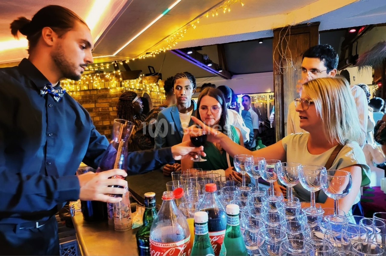 Un barman sert un verre de vin rouge à une femme lors d'un événement dans un bar éclairé par des guirlandes lumineuses.