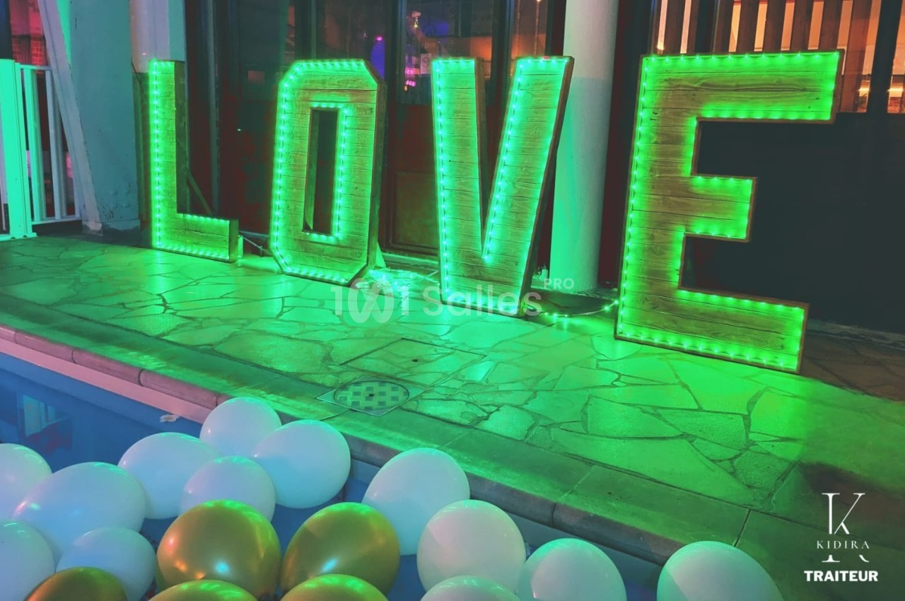 Lettres lumineuses formant le mot ’LOVE’ éclairées en vert, entourées de ballons blancs et dorés près d'une piscine.