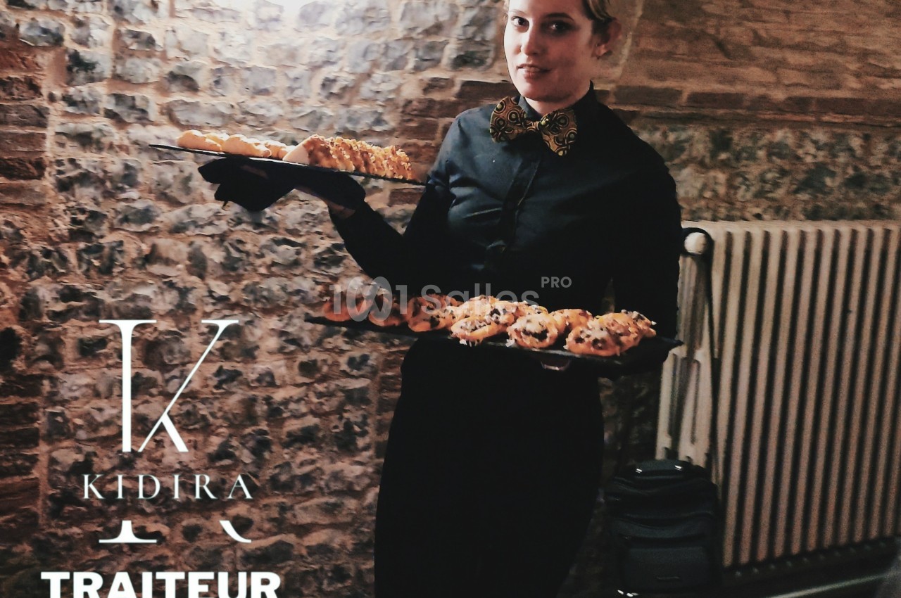 Serveuse en tenue noire portant deux plateaux de viennoiseries dans un cadre en pierre et éclairage tamisé.