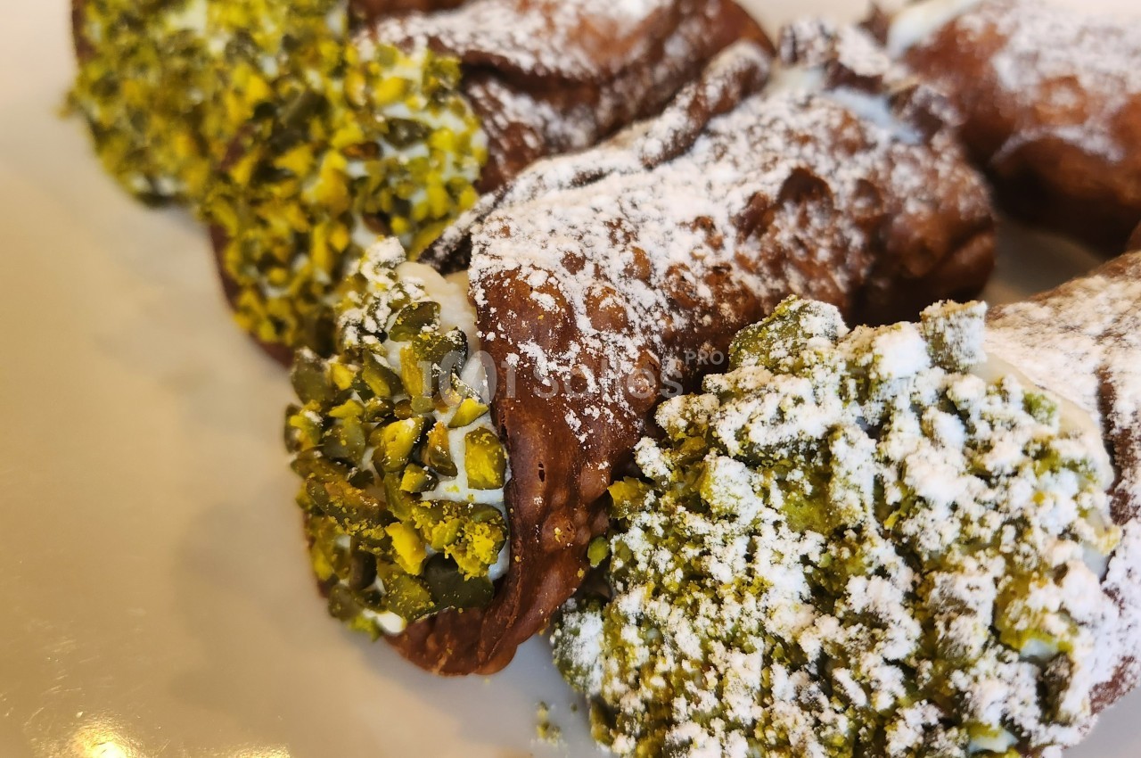 Cannoli garnis de crème, décorés de pistaches concassées et saupoudrés de sucre glace, présentés sur une assiette.