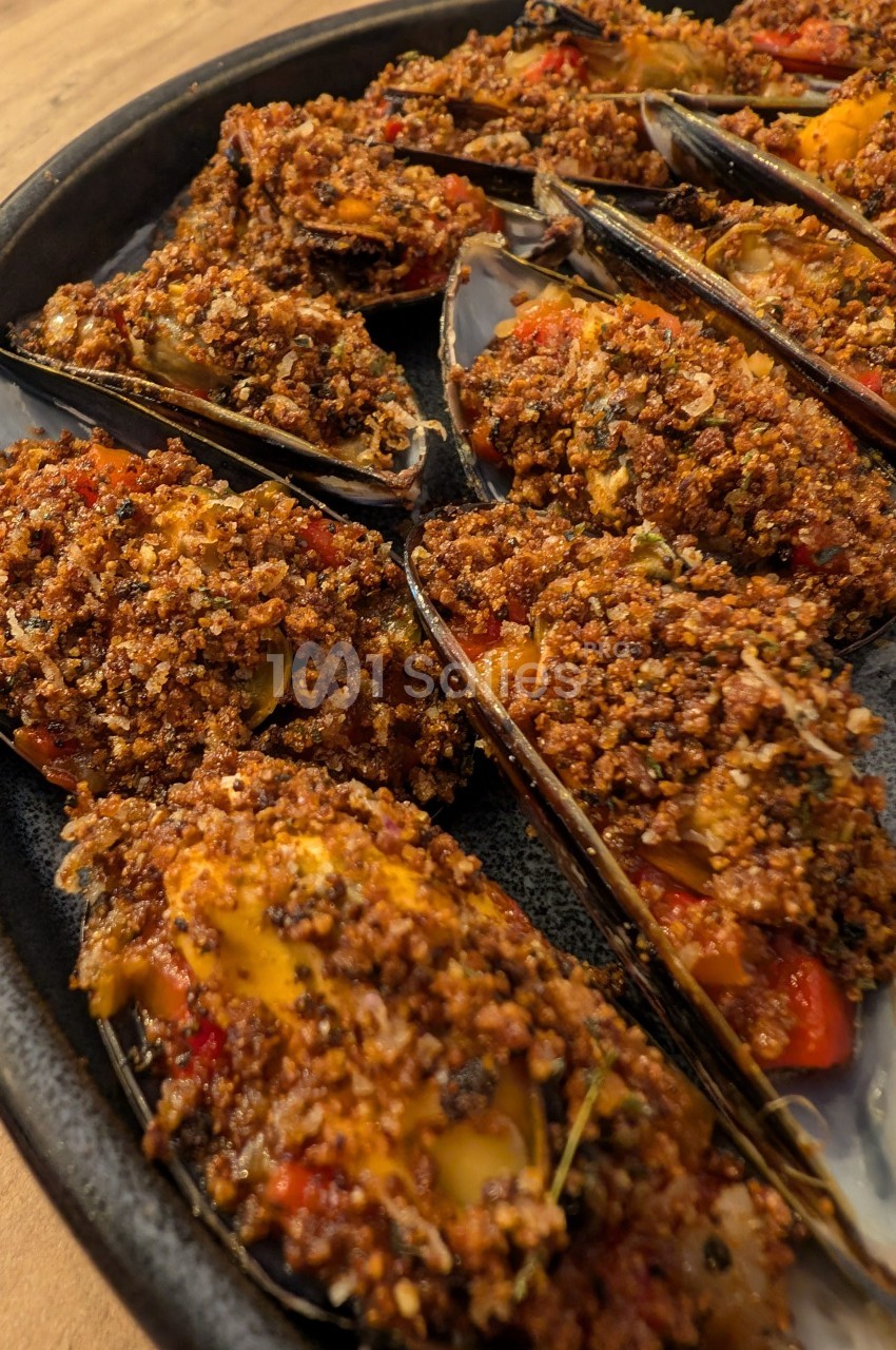 Moules gratinées garnies de chapelure dorée, herbes et morceaux de légumes, servies sur un plat en céramique.