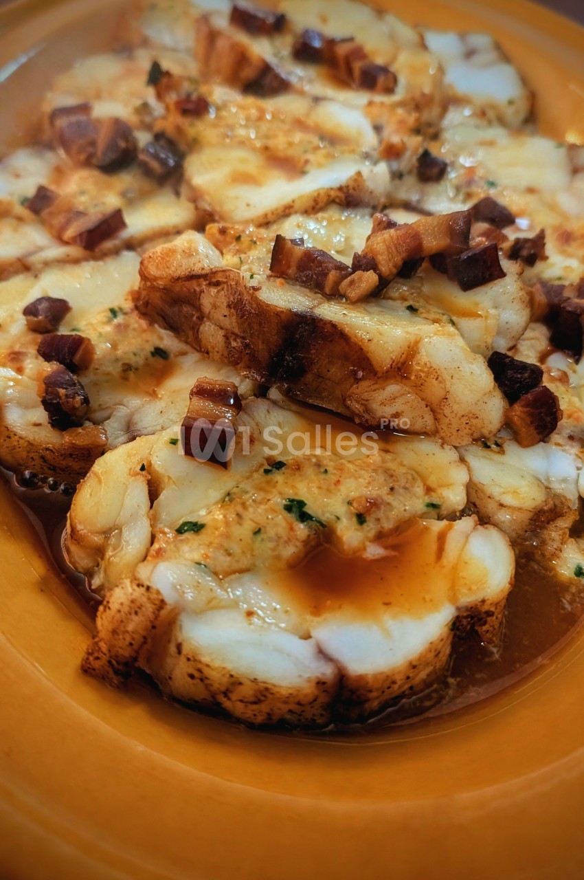 Tranches de lotte servies avec une sauce et des morceaux de lard sur un plat en terre cuite.