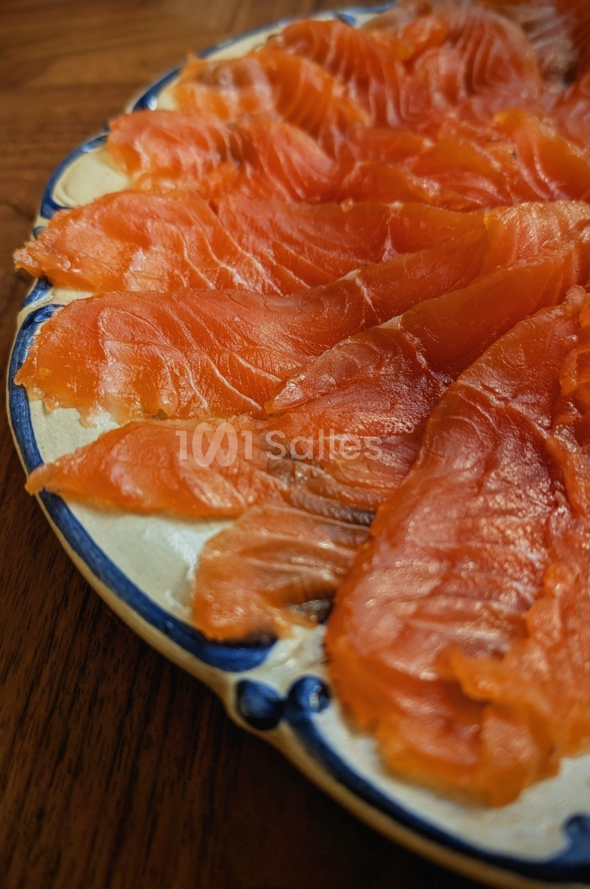 Tranches de saumon fumé disposées en éventail sur une assiette blanche bordée de bleu, posée sur une table en bois.